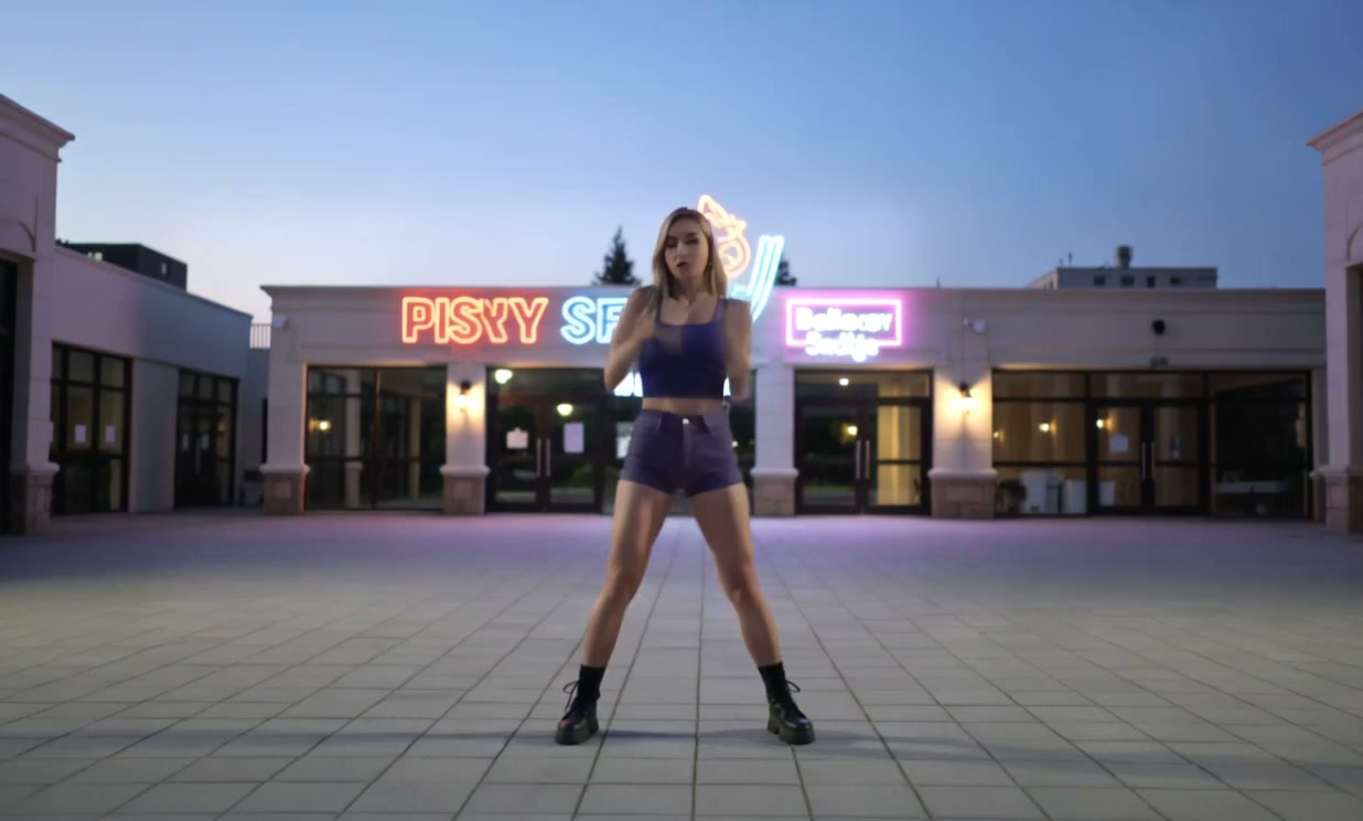 One AI dancer in a purple outfit in a neon-lit plaza at dusk.